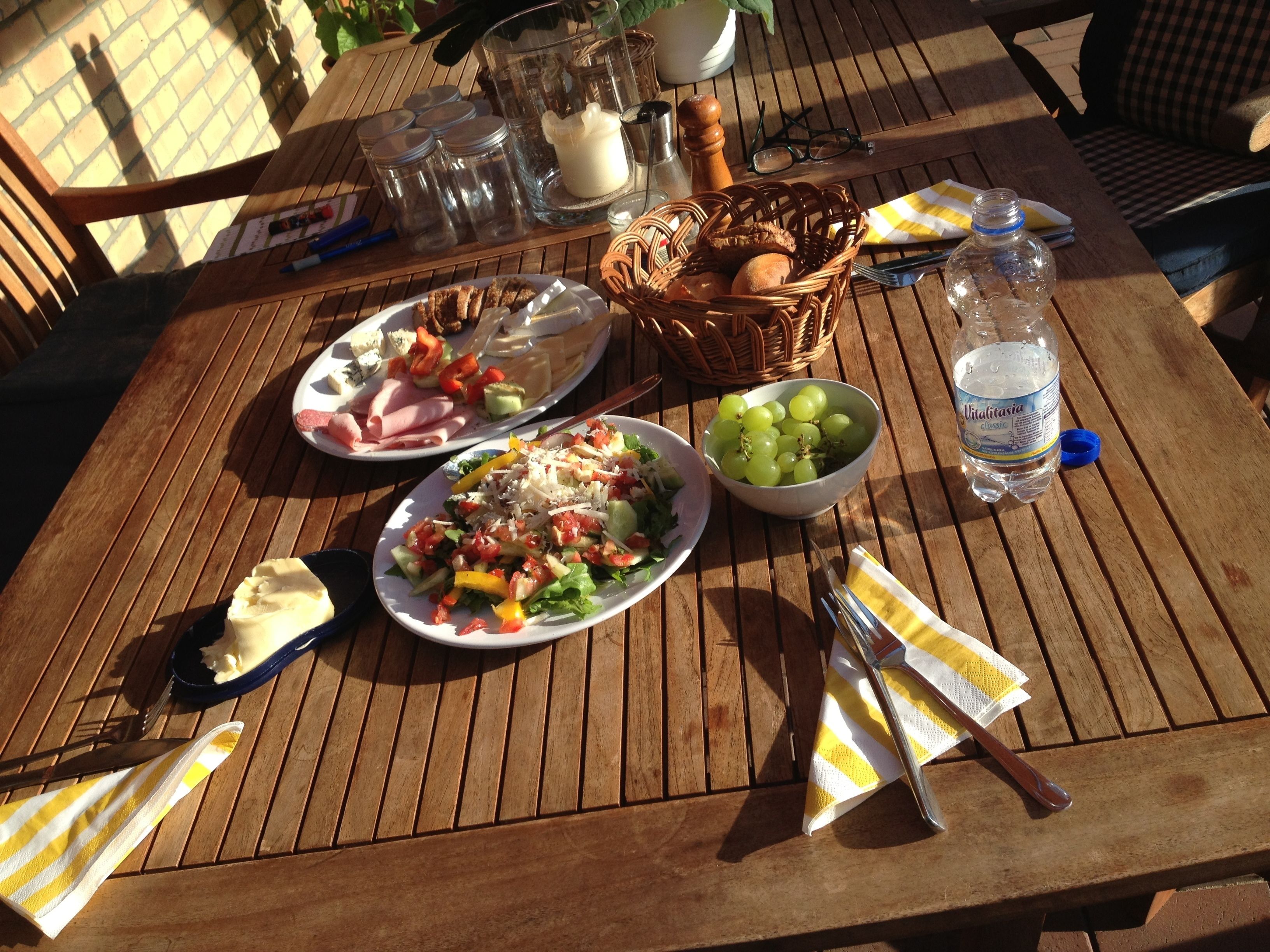 Ein Holztisch mit Tellern voller Essen, einem Brotkorb, einer Schüssel mit Trauben und einer Wasserflasche.