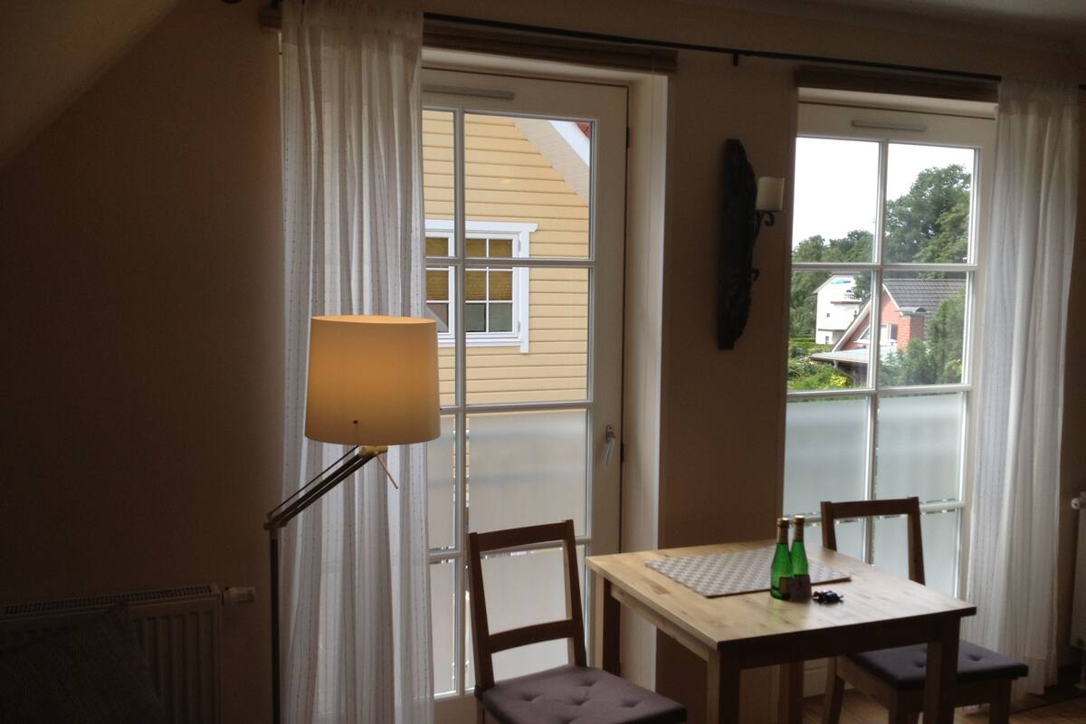A room with a wooden table, two chairs, a lamp, and a view through a large window.