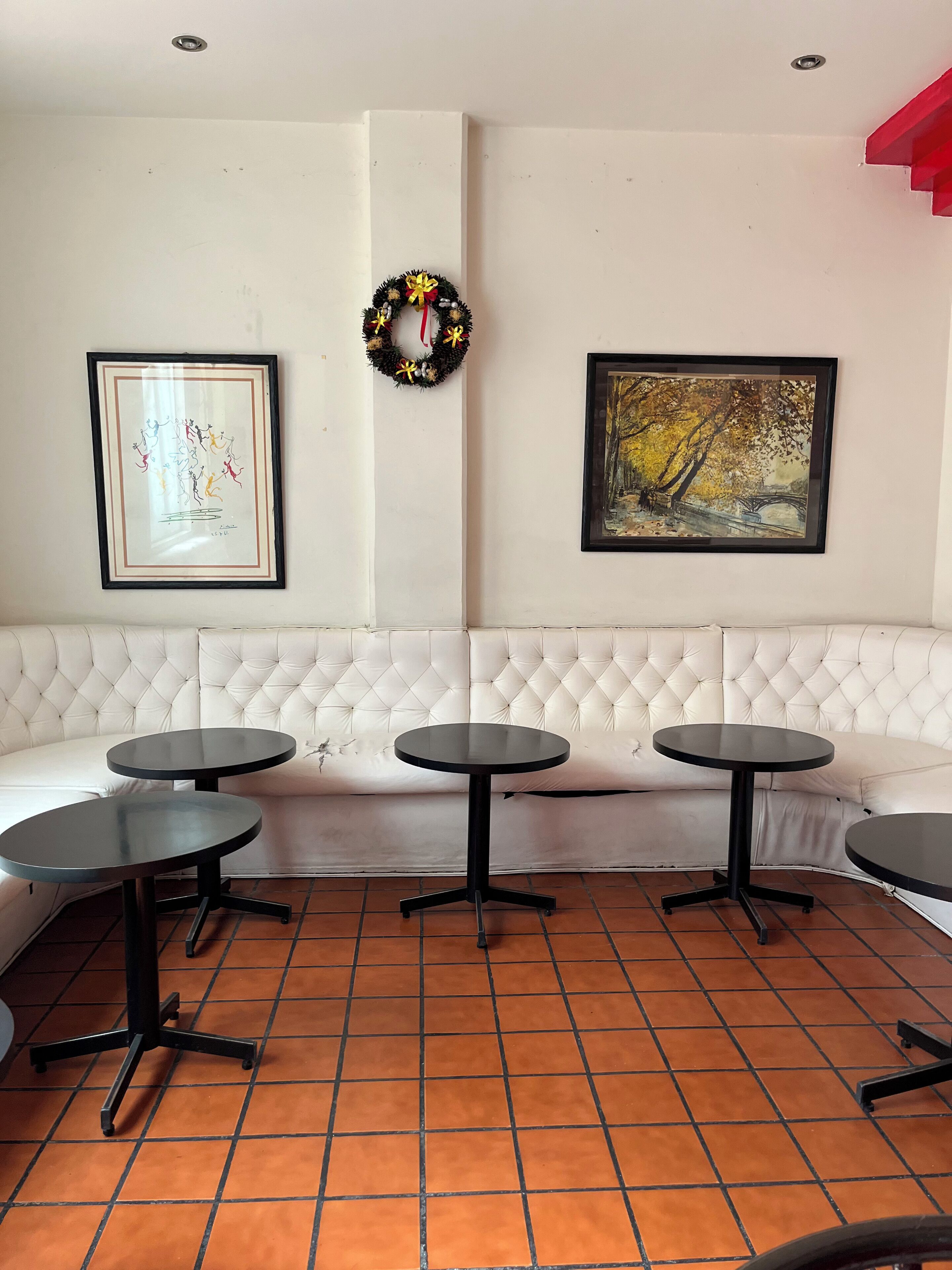 Une pièce avec un banc blanc rembourré, trois tables noires, une œuvre d’art encadrée, une couronne et un sol carrelé.