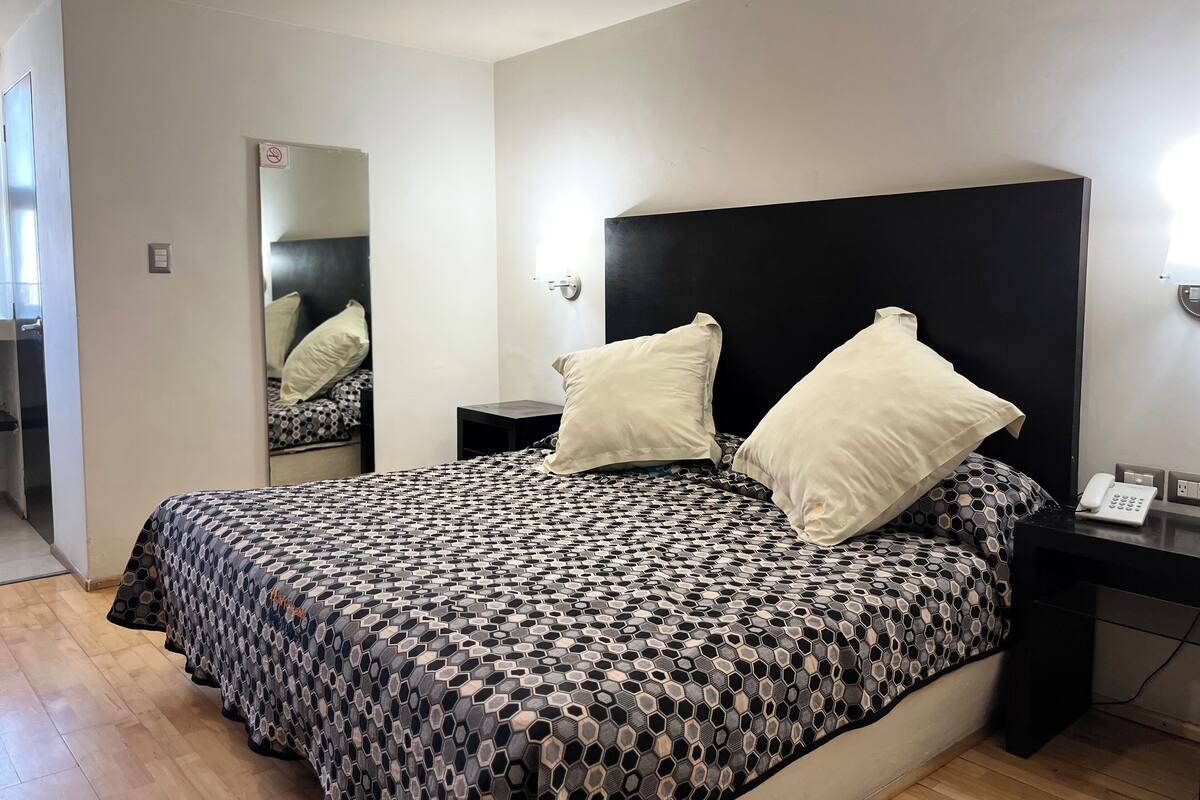 A bedroom with a patterned bedspread, a black headboard, two bedside tables with lamps, and a mirror on the wall.