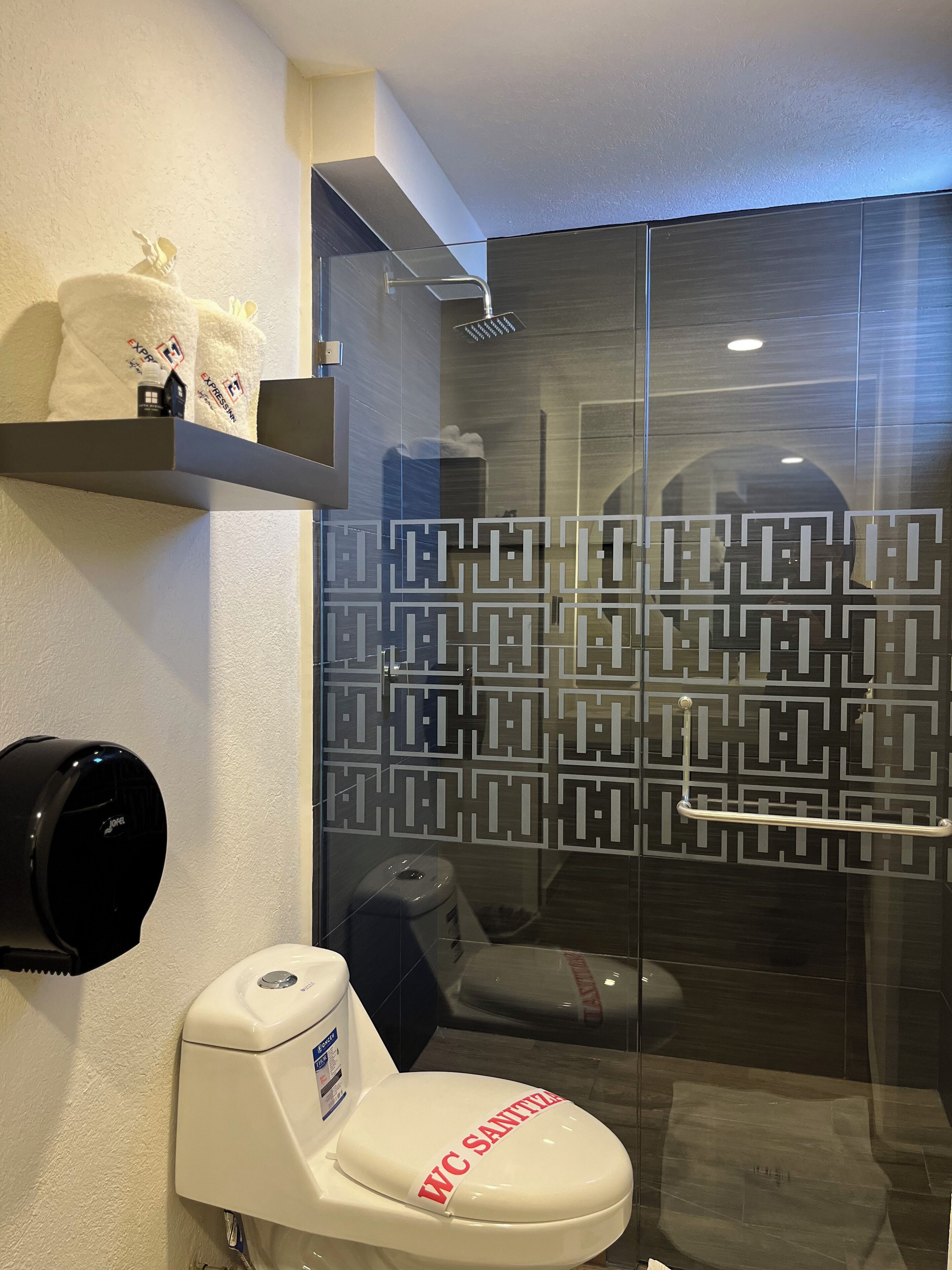 A modern bathroom with a glass shower enclosure, a toilet with a "WC SANITARY" label, a towel dispenser, and a shelf with toiletries.