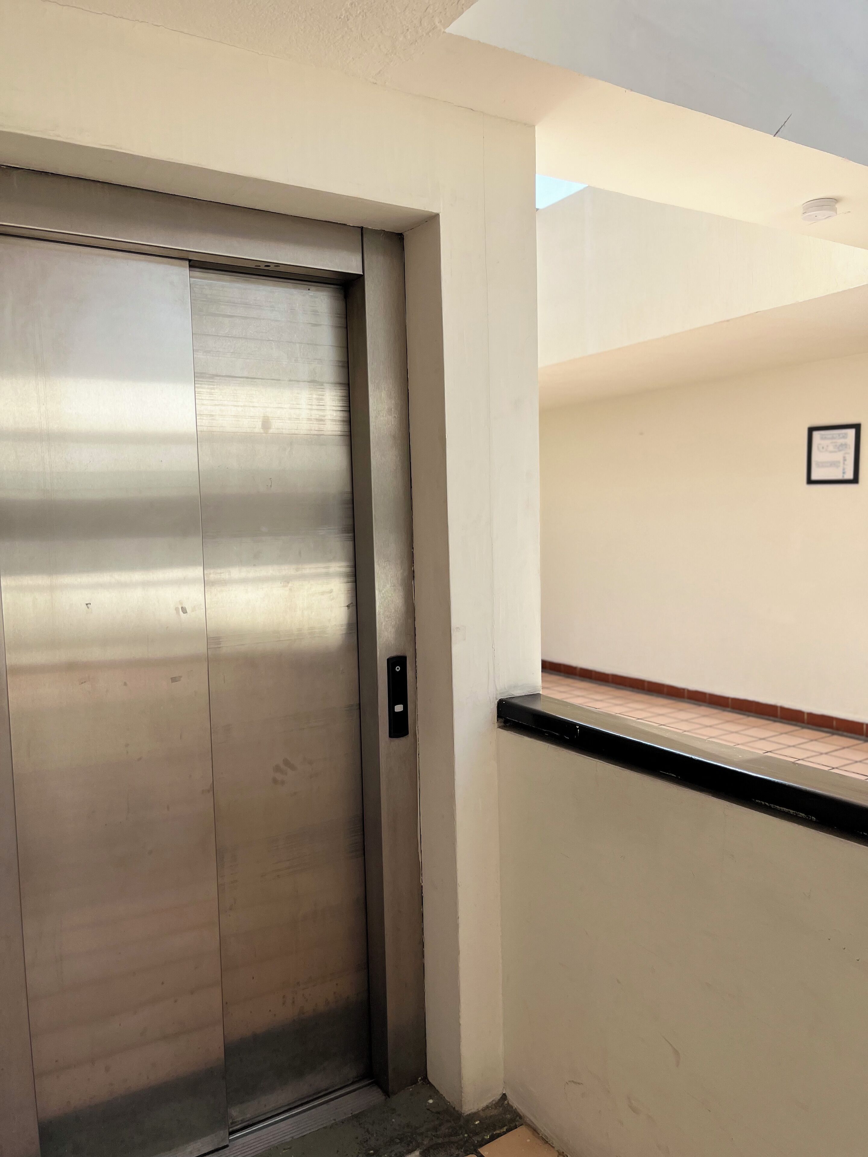 A metallic elevator door in a hallway with a tiled floor and a white wall with a small framed picture.