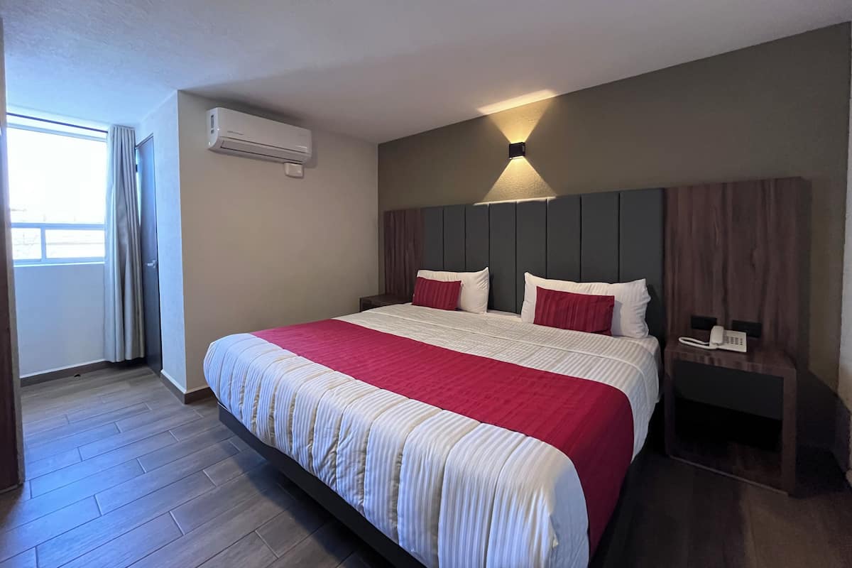 A hotel room with a large bed, a bedside table with a phone, a wall-mounted air conditioner, and a window with curtains.