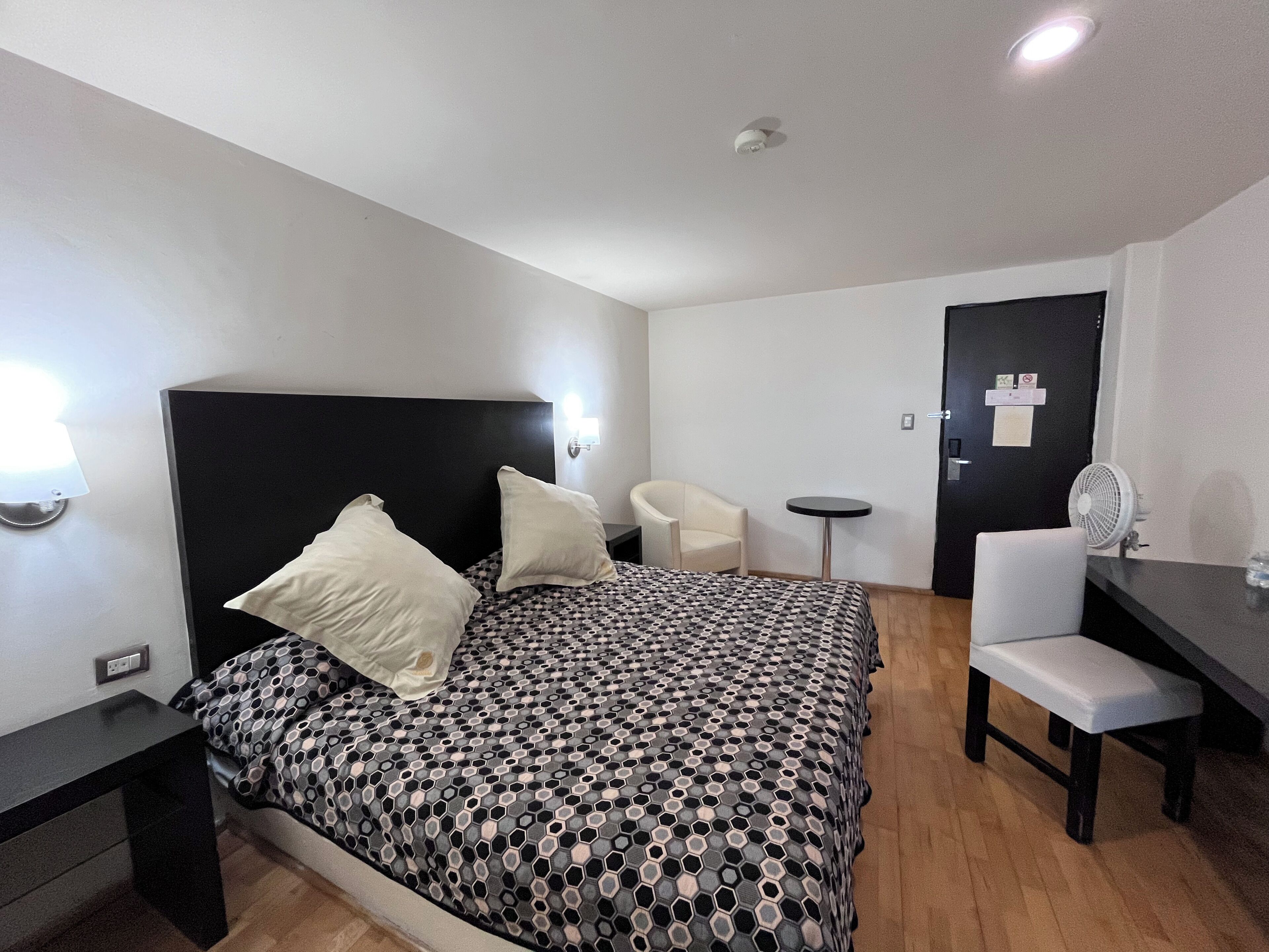 A hotel room with a bed, a desk, a chair, a small table, and a wall-mounted fan.