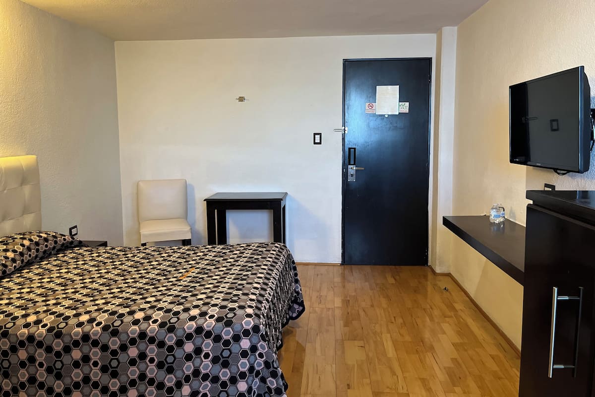 A hotel room with a bed, a desk, a chair, a television, and a door with a notice.