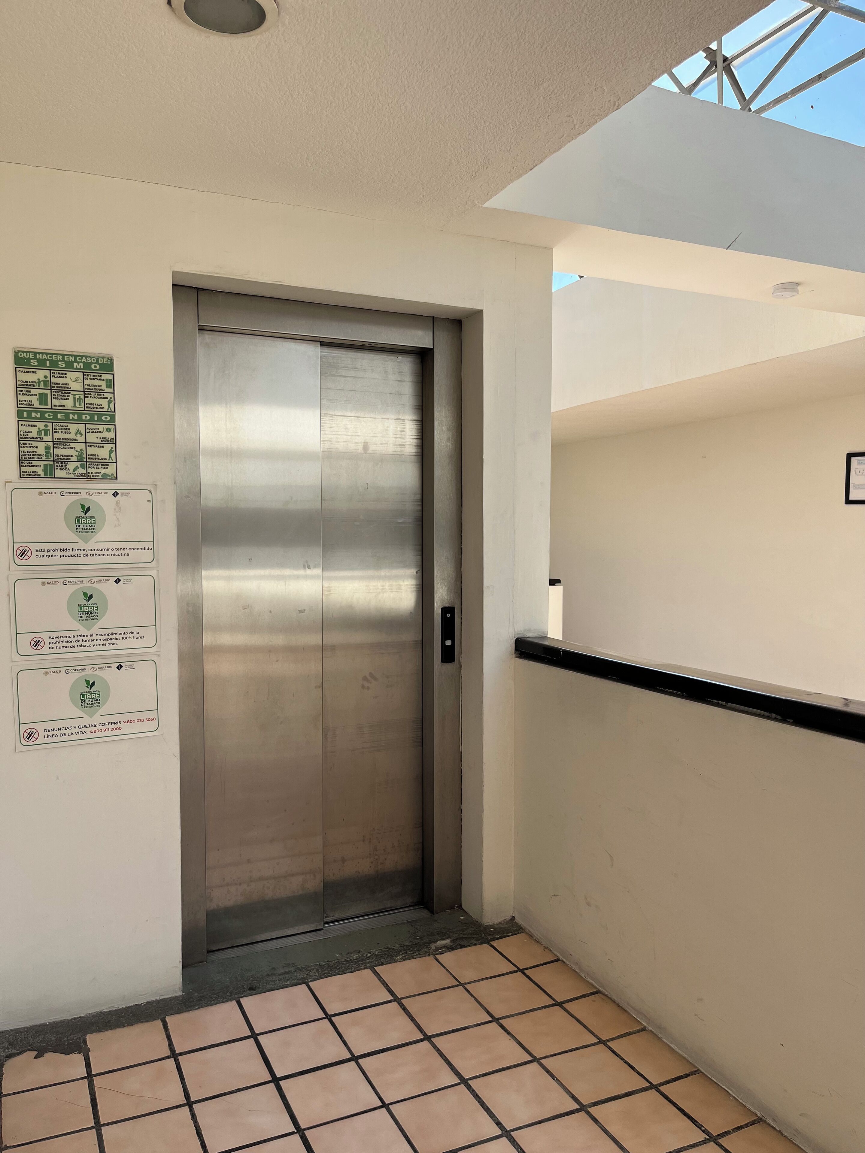 Um elevador com porta metálica, piso de azulejos e parede com cartazes informativos.