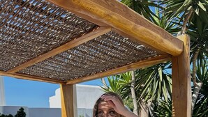 A person sitting on a cushioned outdoor lounge chair under a wooden pergola, with a view of a white building and greenery in the background. - Bellissimo Resort (Mykonos)