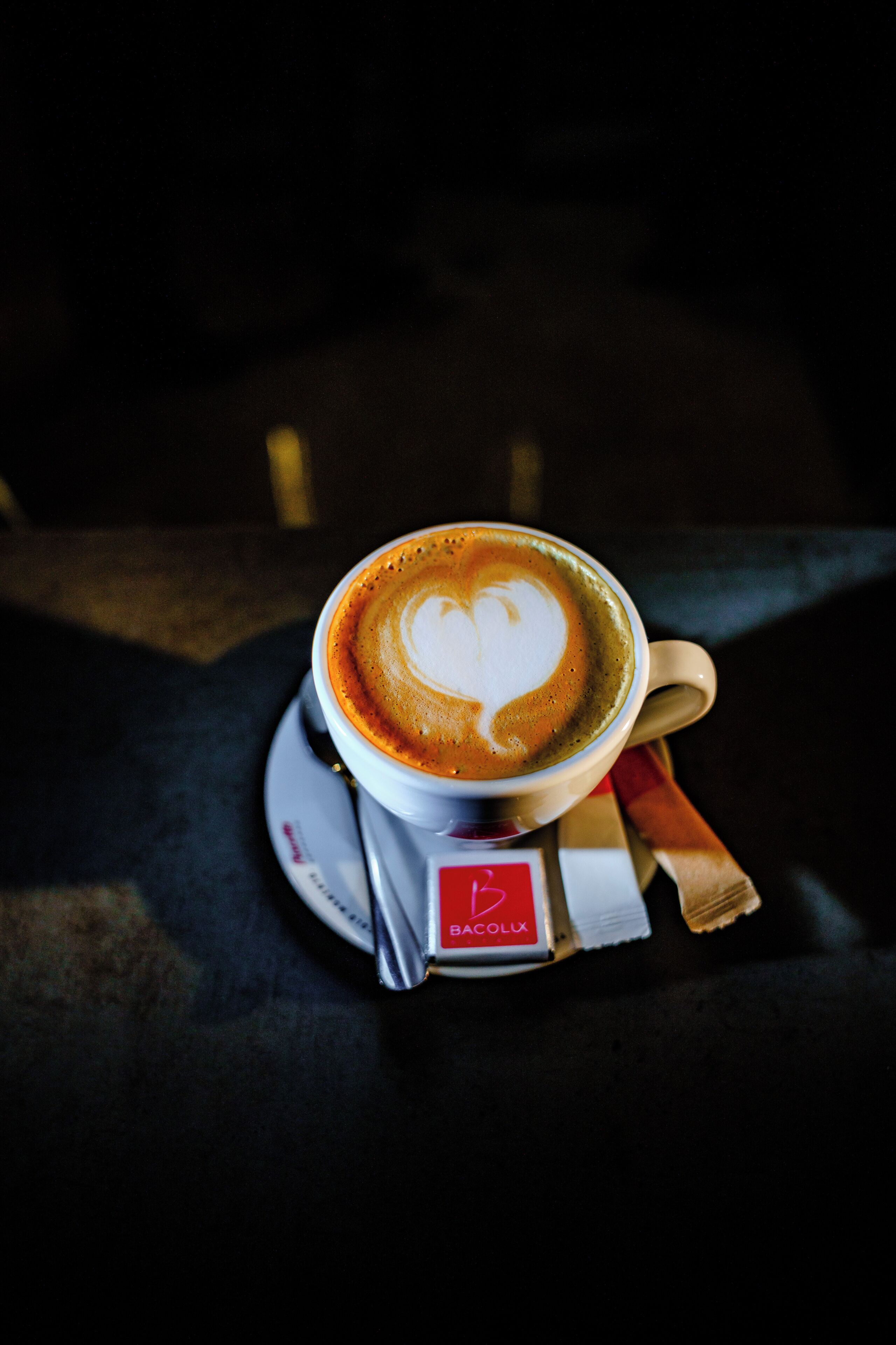 A cup of coffee with a heart-shaped latte art on a saucer, placed on a table with a napkin and a small box labeled 'BACOLUX.'