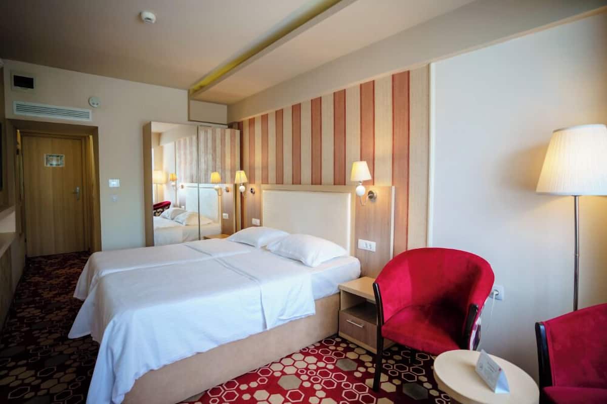 Una habitació d'hotel amb un llit, una cadira vermella, una taula petita, un mirall i una làmpada.