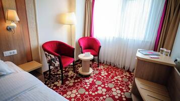 Quarto de hotel com uma cama, duas cadeiras vermelhas, uma mesa redonda pequena, uma escrivaninha com abajur e uma janela com cortinas.