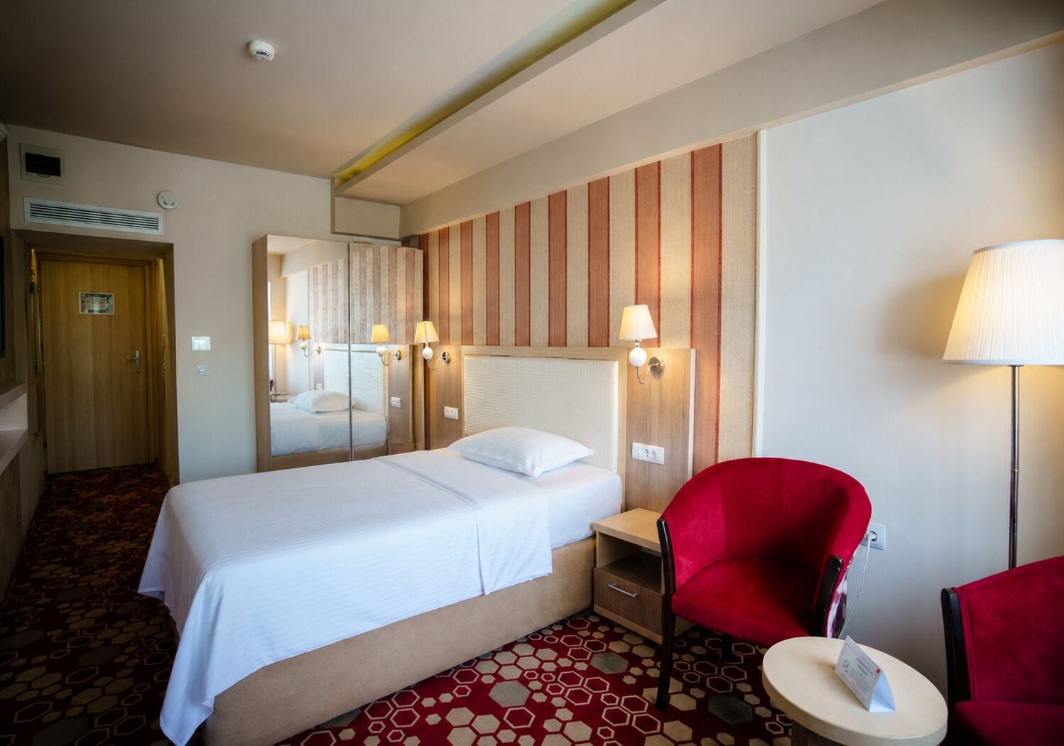 A hotel room with a bed, a red chair, a small table, a lamp, a mirror, and a door.