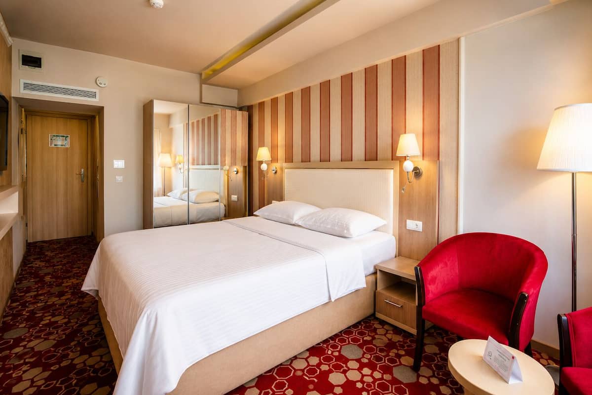 Habitació d'hotel amb un llit gran, una butaca vermella, una taula petita i un mirall.