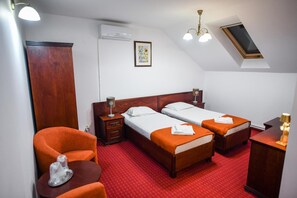 A hotel room with two beds, a wooden wardrobe, a small table with a chair, and a ceiling fan.