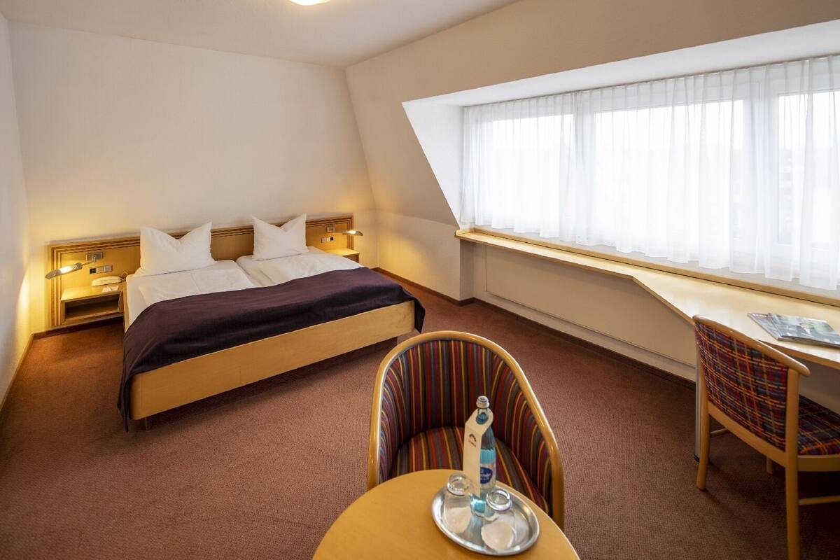 Ein Hotelzimmer mit einem Bett, einem Schreibtisch, einem Stuhl und einem Fenster mit durchsichtigen Vorhängen.
