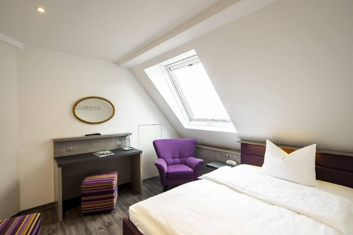 Ein modernes Hotelzimmer mit Dachfenster, einem Schreibtisch, einem violetten Sessel und einem Bett mit weißer Bettwäsche.