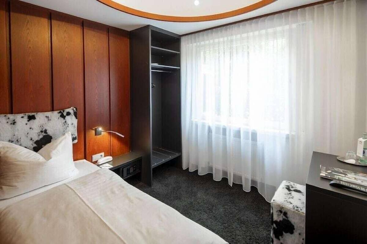 Ein Hotelzimmer mit Bett, Nachttisch, Kleiderschrank, Schreibtisch und einem fenster mit Vorhängen.