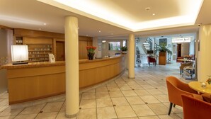 A hotel reception area with a curved reception desk, a coffee machine, and a potted plant. There are chairs and a table in the foreground, and a staircase leading to an upper level. - Hotel Gloria (Stuttgart)