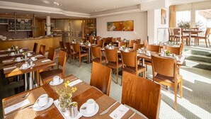 A hotel dining area with multiple tables and chairs, a kitchen in the background, and a large window providing natural light. - Hotel Gloria (Stuttgart)