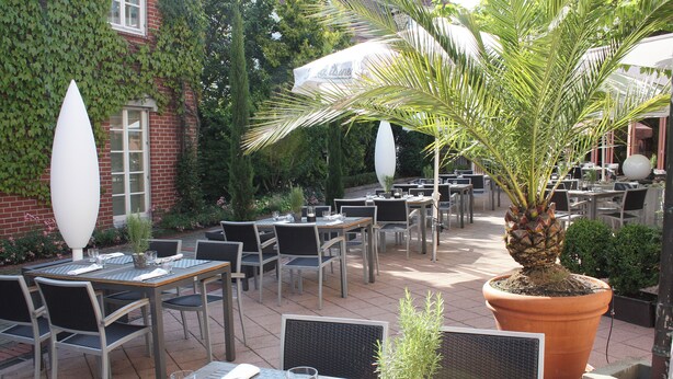 An outdoor patio with tables and chairs, surrounded by greenery and a brick building. - Hotel Gloria (Stuttgart)