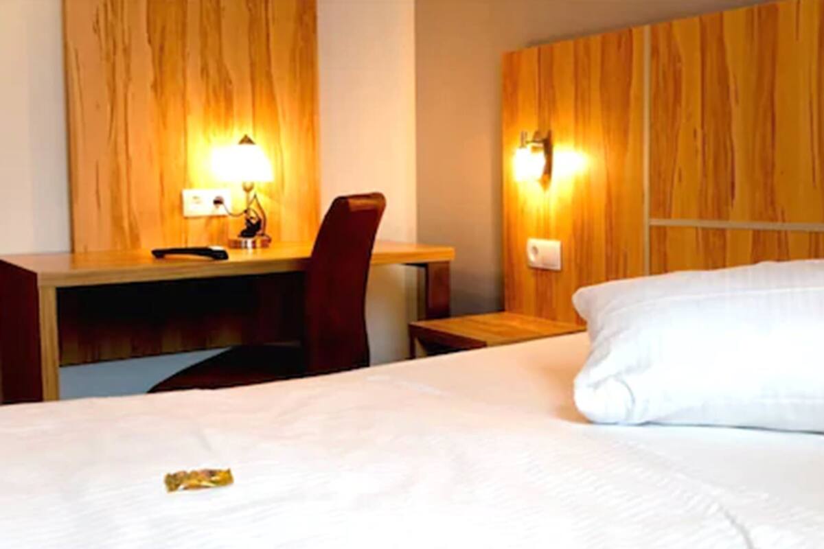 A hotel room with a wooden headboard, a desk with a lamp, a chair, and a neatly made bed.