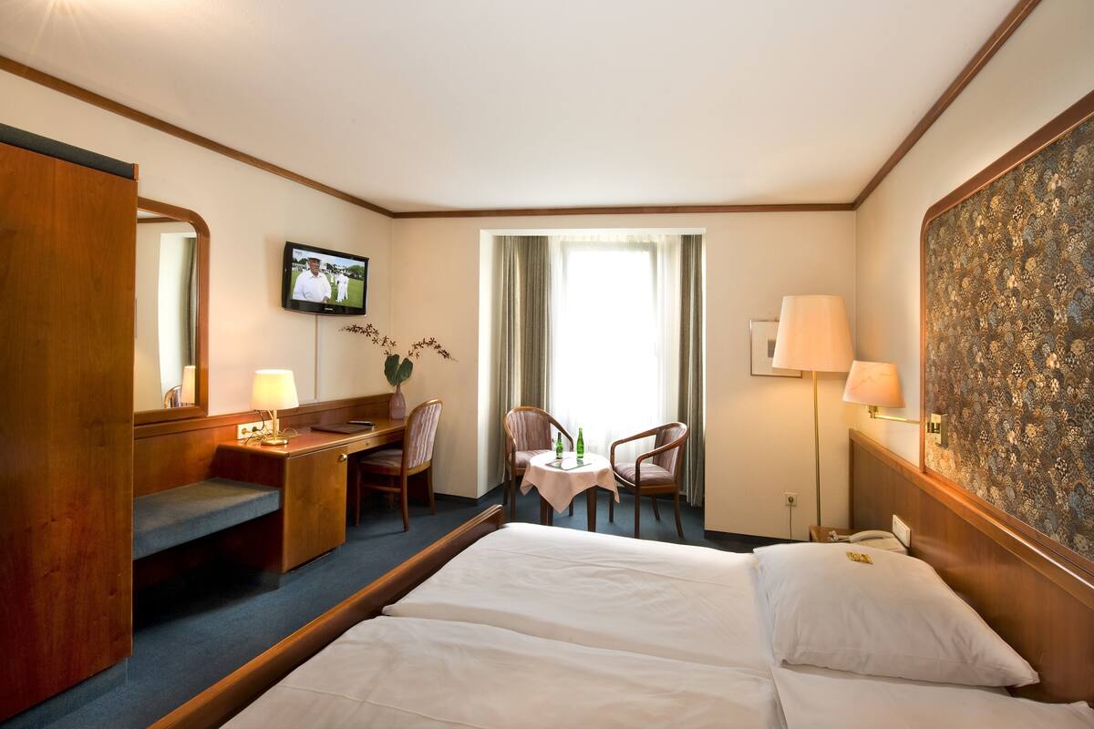 A hotel room with a bed, a desk with a chair, a small table with two chairs, a TV, and a window with curtains.