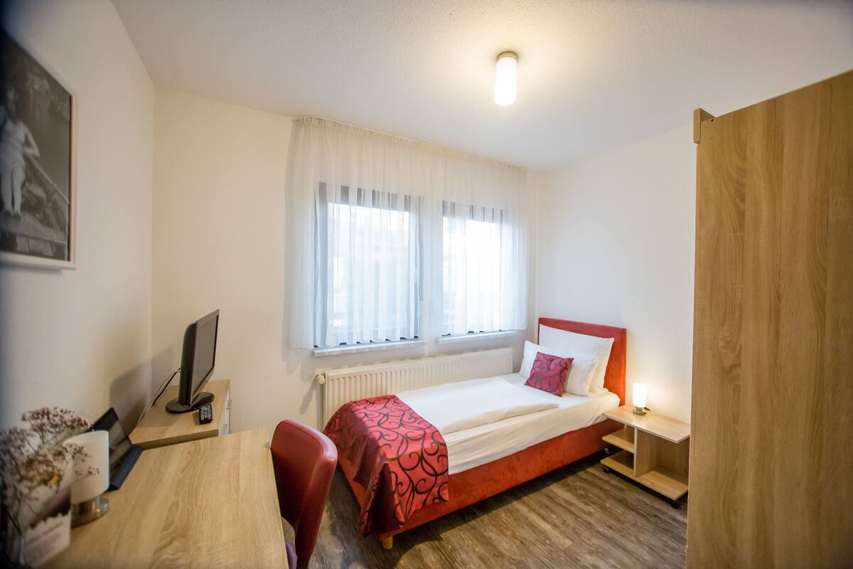 Un dormitorio con cama, escritorio, silla, televisor y ventana con cortinas.