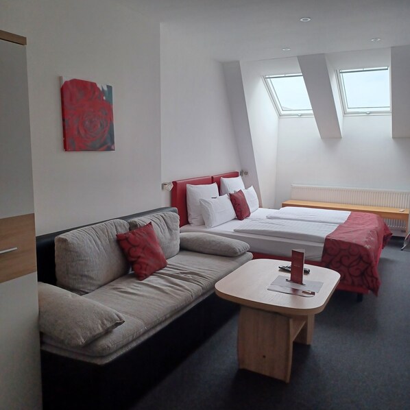 Une chambre d’hôtel moderne avec un canapé, un lit avec une literie rouge et blanche, une petite table en bois et un tableau abstrait rouge accroché au mur.