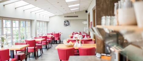 Un restaurante con sillas rojas y mesas de madera, ventanas amplias e iluminación empotrada en el techo.