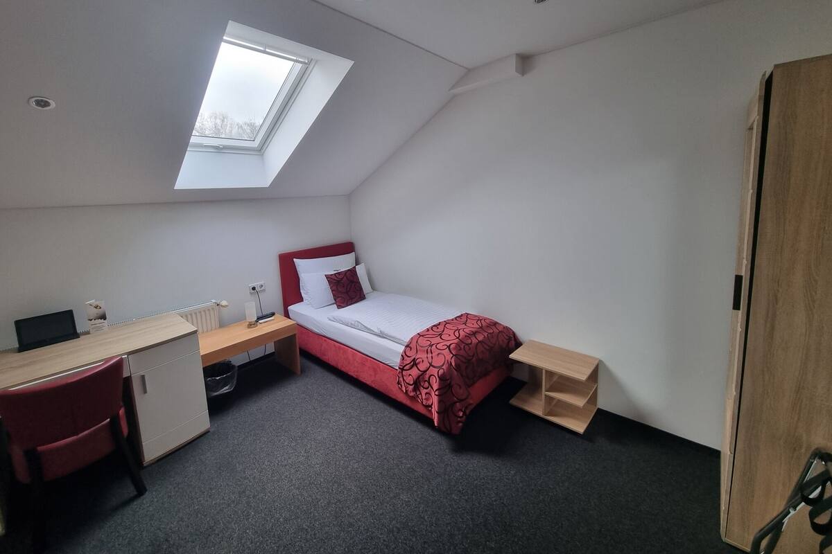 Habitación pequeña con cama, escritorio y silla. Hay un tragaluz sobre la cama y un radiador en la pared.