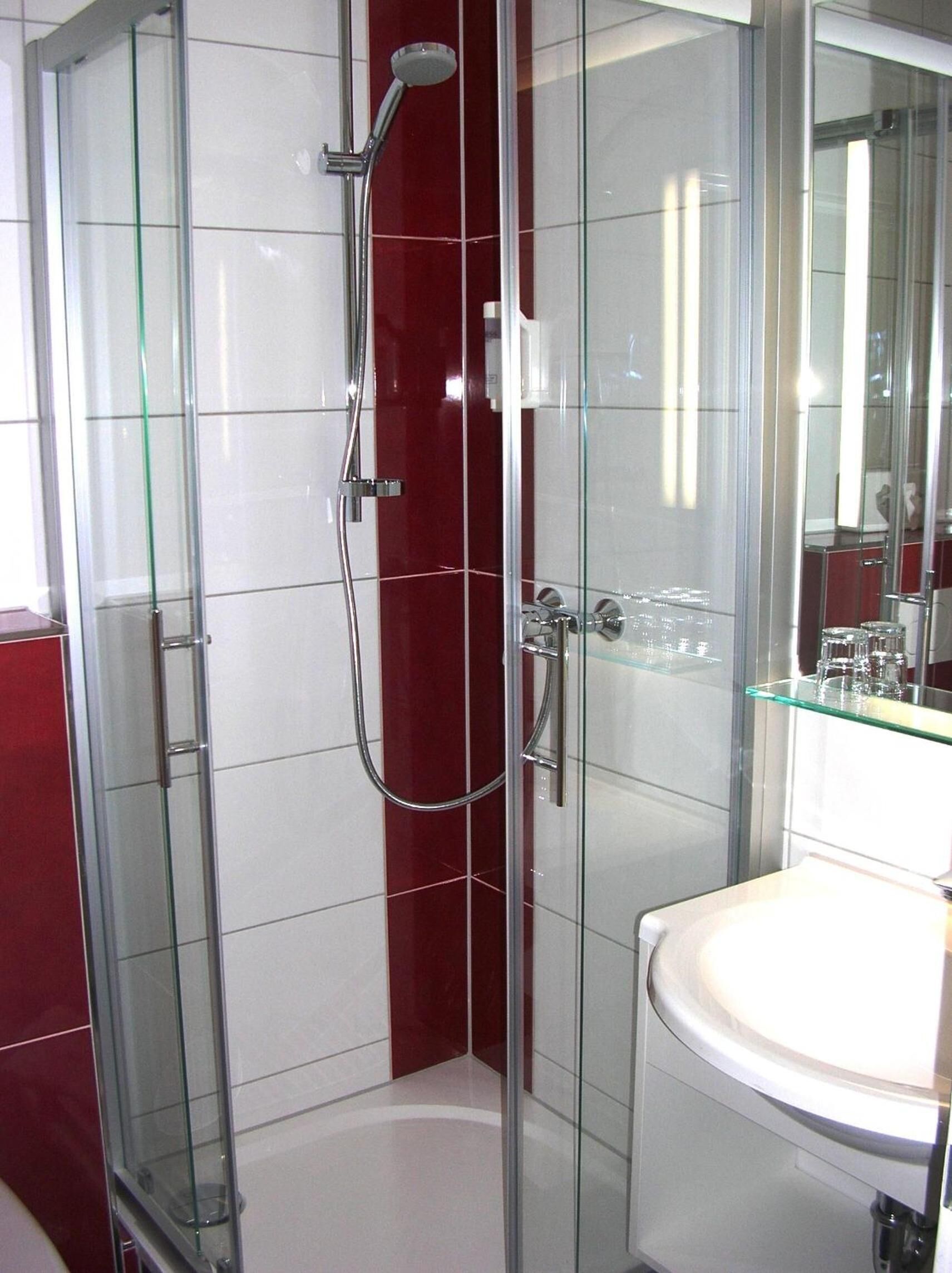 Un baño moderno con cabina de ducha de vidrio, un lavamanos blanco y una pared de acento roja.