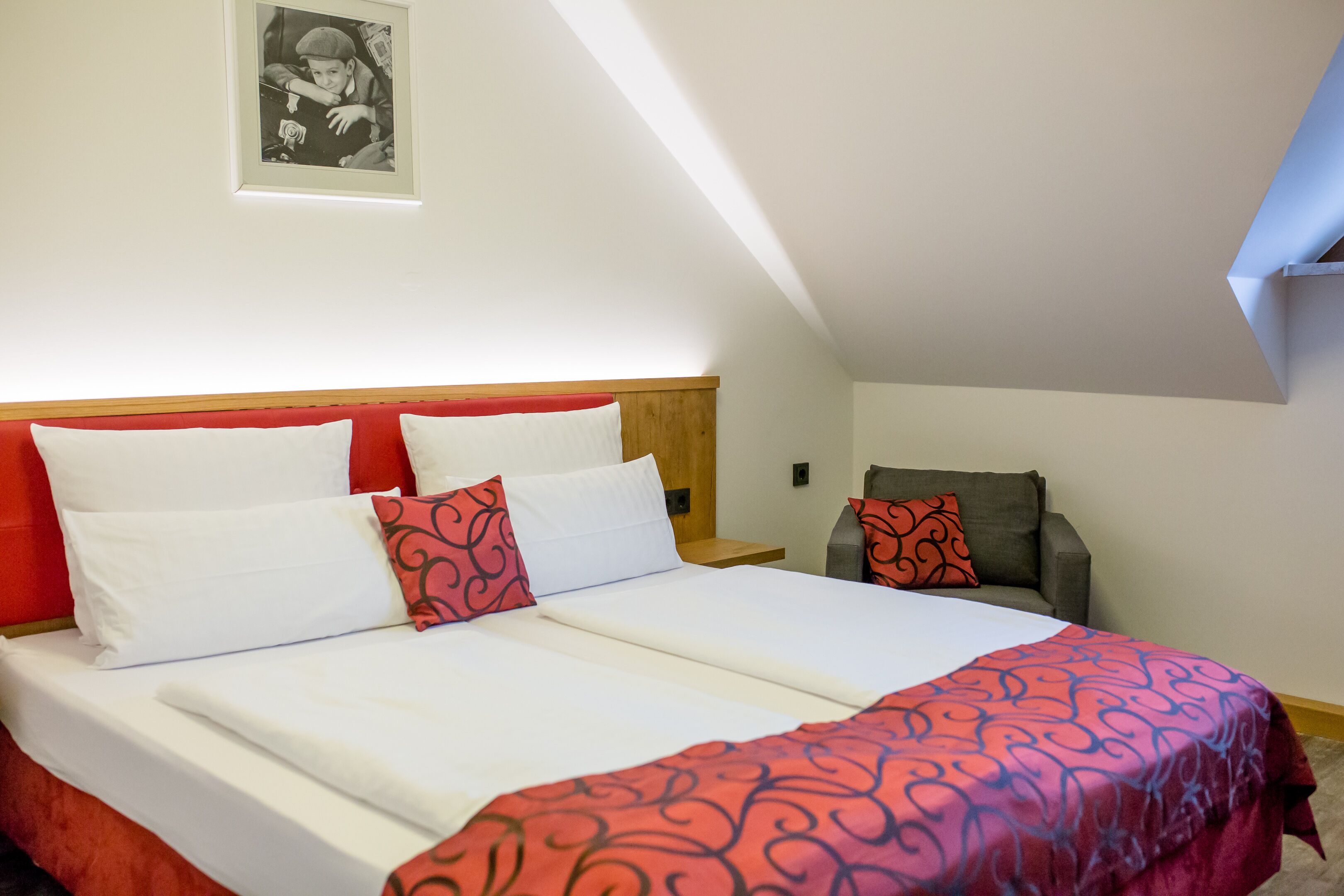A double bed with white and red bedding, a wooden headboard, and a small bedside table.