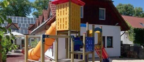Un parque infantil con un tobogán y estructuras para escalar, frente a una casa con techo rojo y paneles solares.