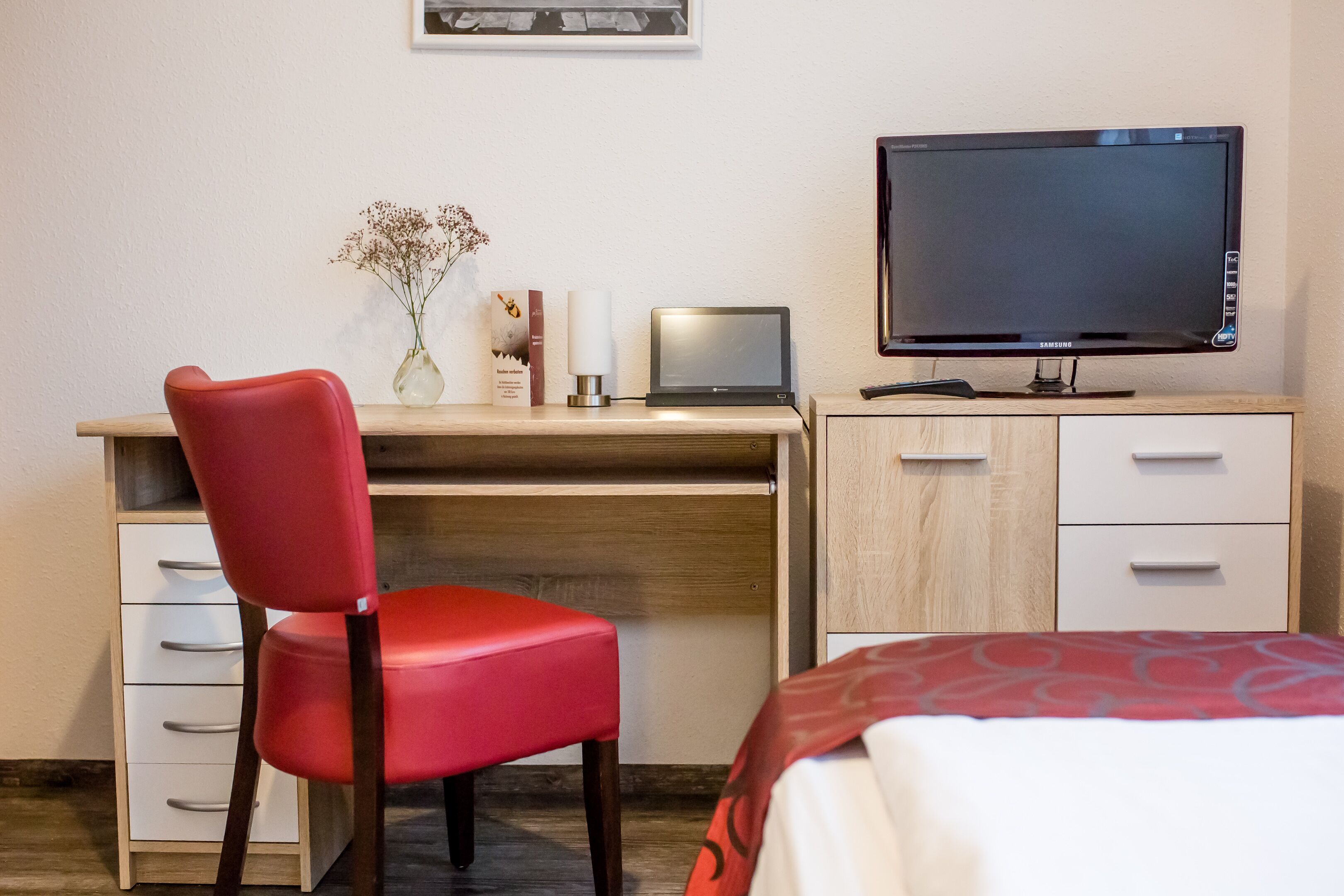 Quarto de hotel com uma secretária de madeira, uma cadeira vermelha, uma televisão de tela plana, uma cama com colcha estampada em vermelho e branco, e um vaso com flores na secretária.