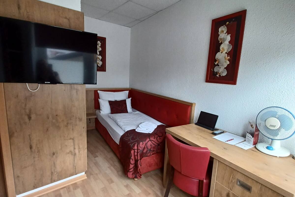 Una habitación compacta con cama, escritorio, silla, televisor y ventilador de pared.