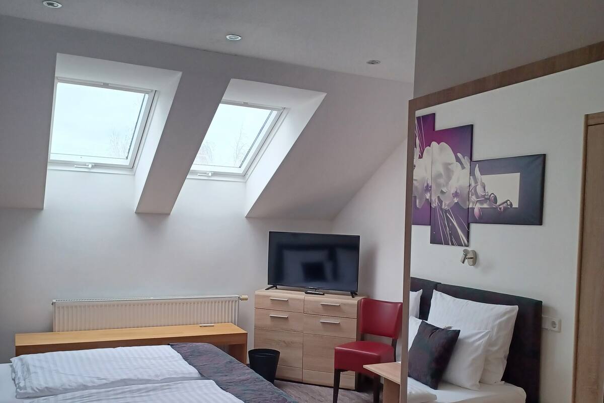 Habitación de hotel moderna con una cama, un escritorio, un televisor y una silla roja. Hay tragaluces y un cuadro en la pared.