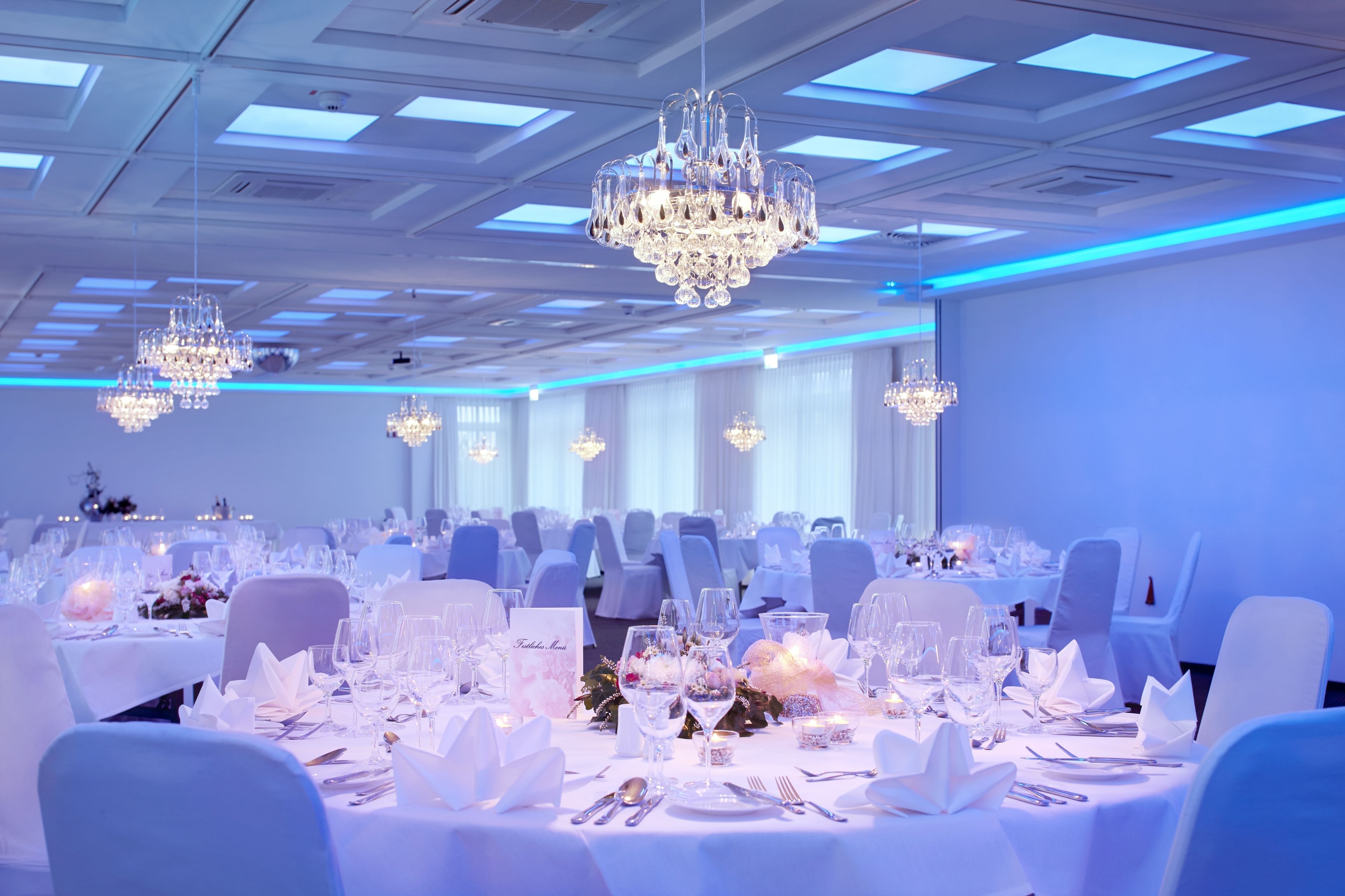 A banquet hall with multiple tables set for a formal event, featuring chandeliers and white tablecloths.