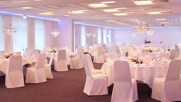 Une salle de banquet avec des tables rondes, des chaises blanches, des lustres et un sol recouvert de moquette.