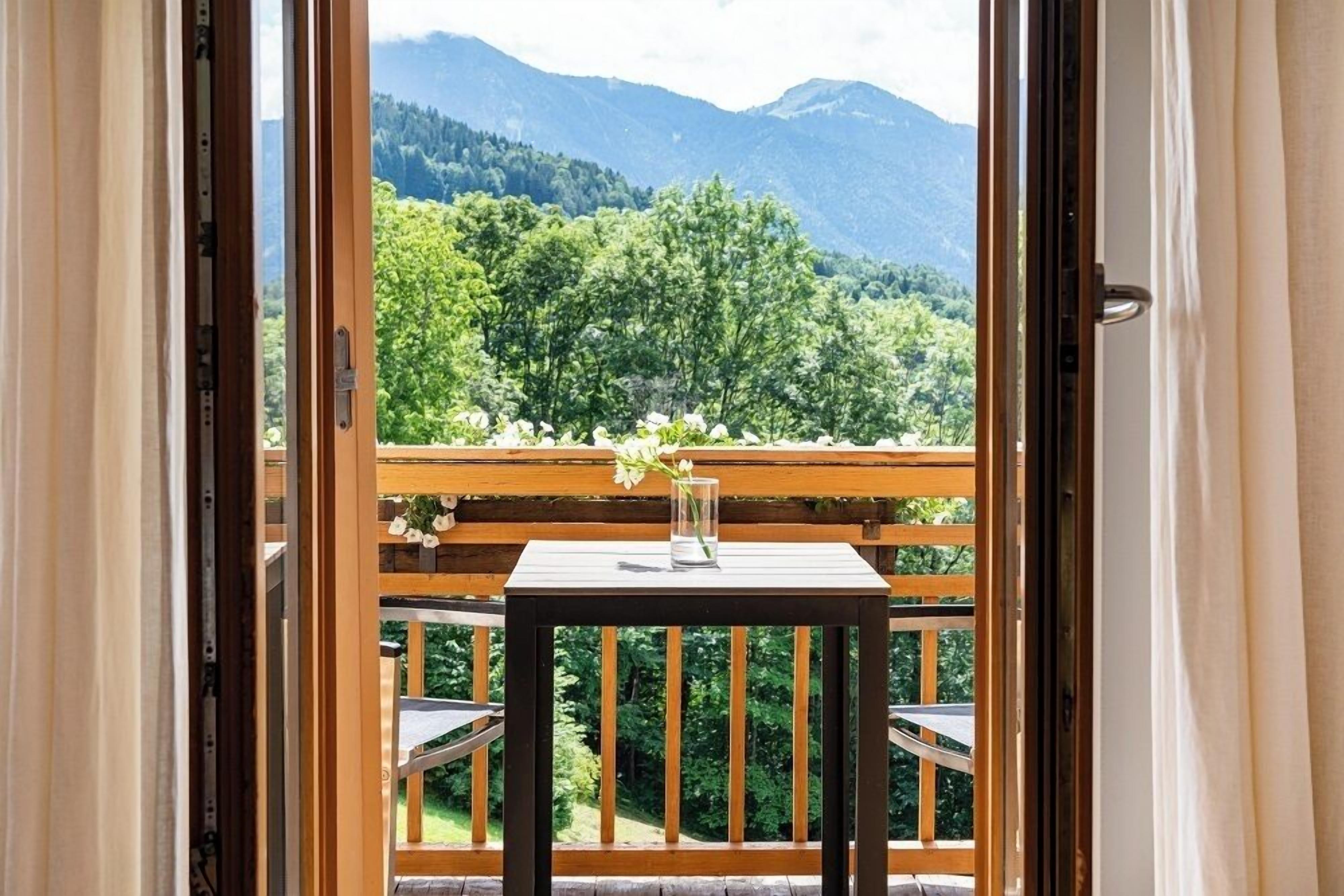 Un balcone con tavolo e sedie in legno, un vaso con fiori e vista su una catena montuosa ricoperta di boschi.
