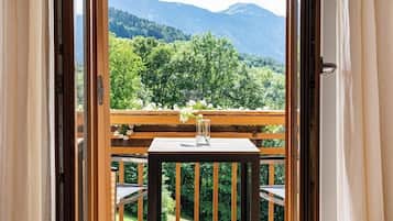 Un balcone con tavolo e sedie in legno, un vaso con fiori e vista su una catena montuosa ricoperta di boschi.