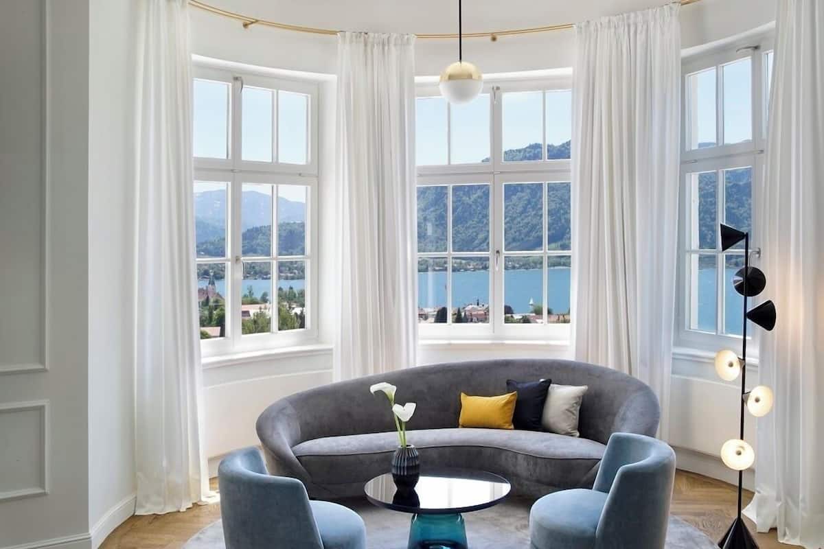 A modern living room with a curved grey sofa, two armchairs, a round coffee table, and a vase with flowers. Large windows offer a scenic view of mountains and a lake.