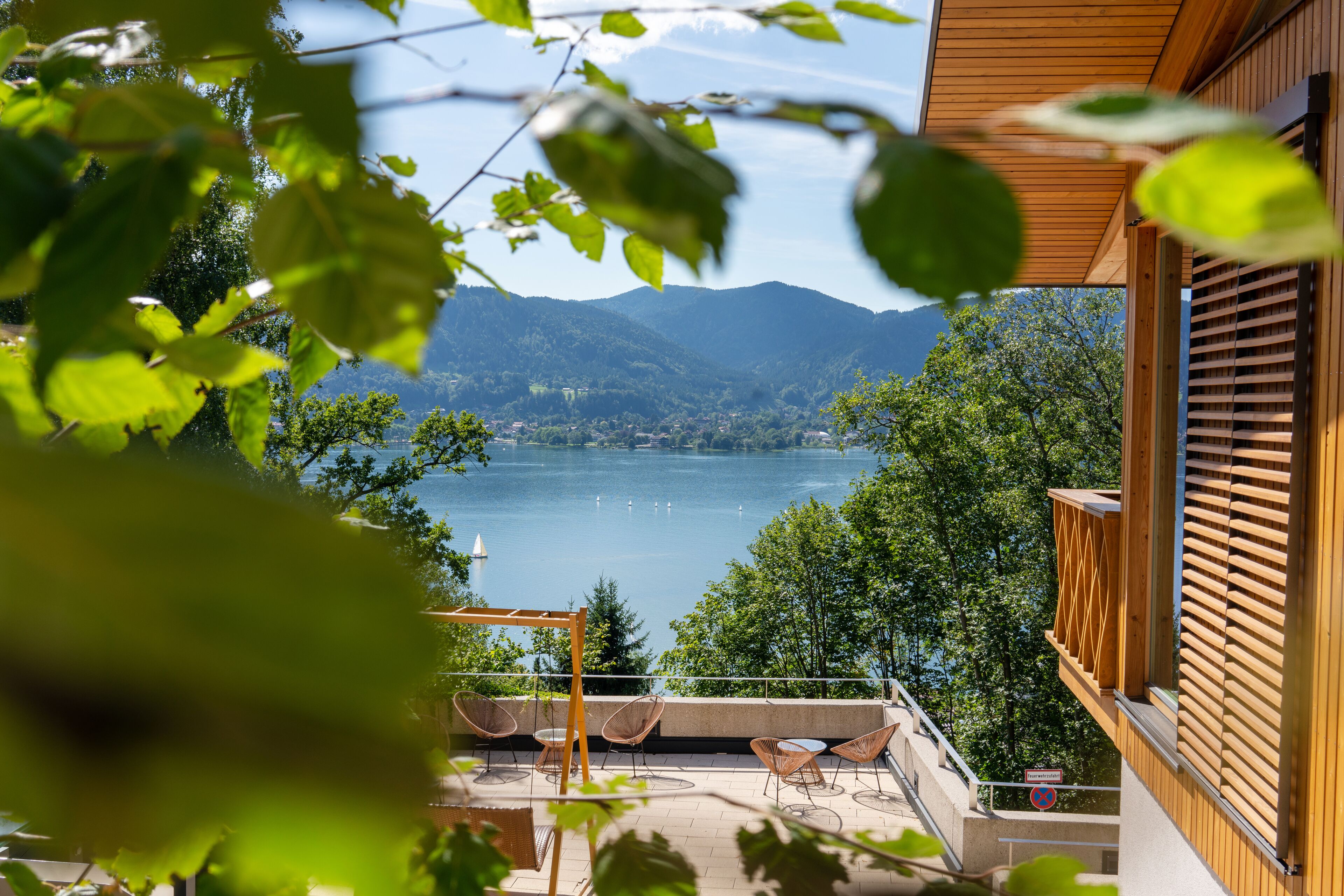 Un balcón con vistas a un lago y montañas, barandas de madera y mobiliario exterior.