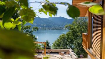 Un balcón con vistas a un lago y montañas, barandas de madera y mobiliario exterior.
