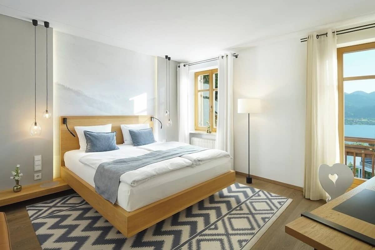 A modern hotel room with a bed, bedside table, lamp, and a view of the outdoors.