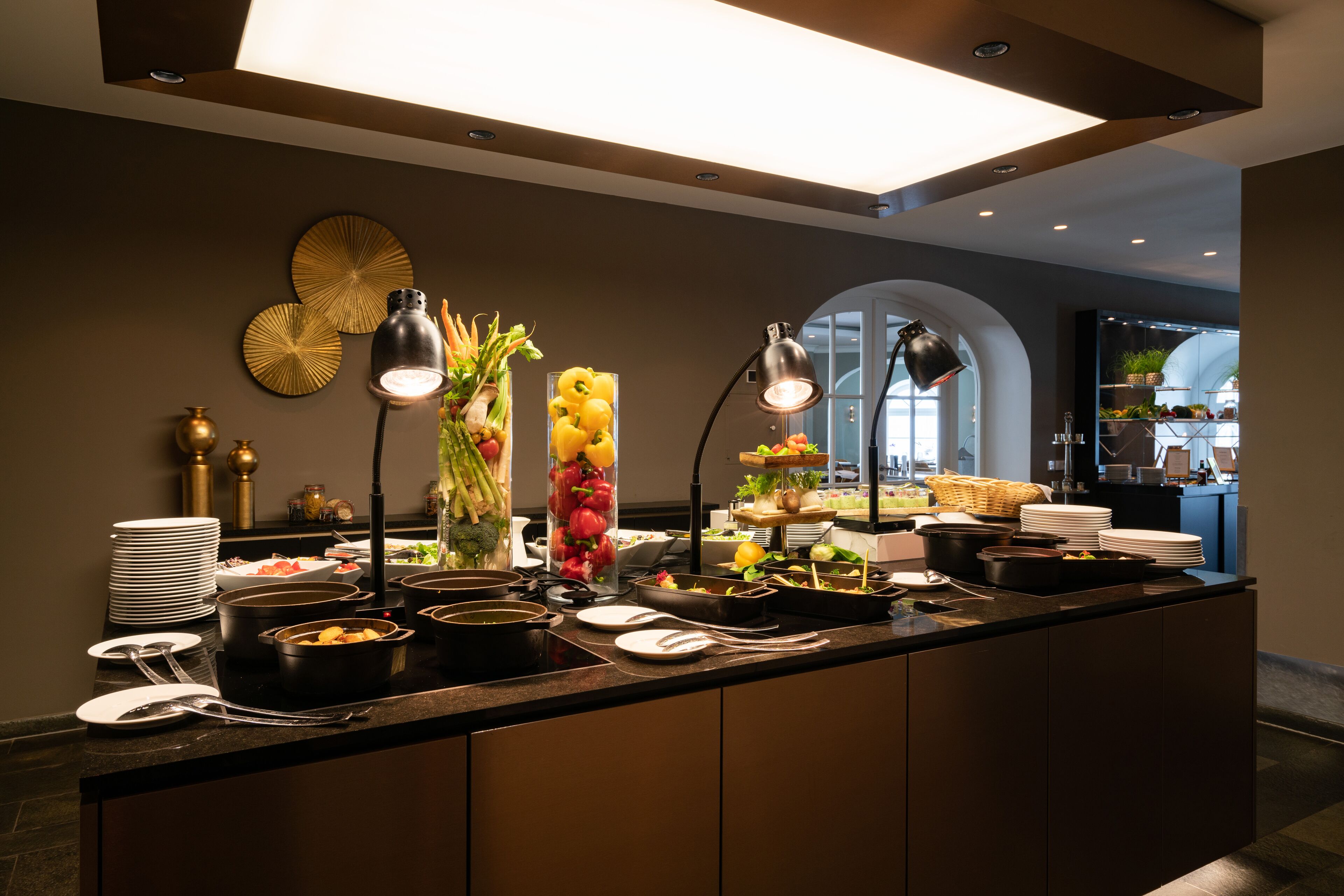Montaje de bufé con diversos platos, una pared decorativa y vista al área de cocina.