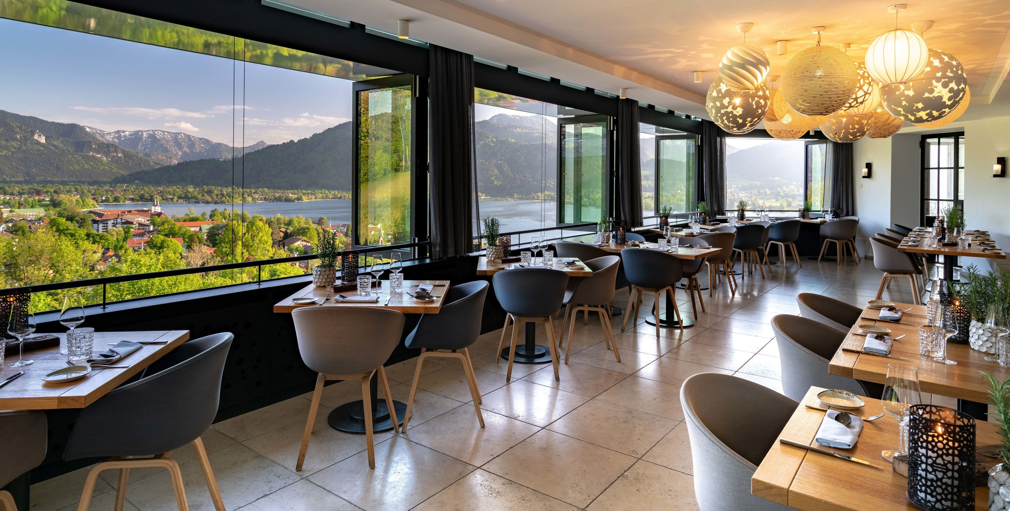 Ein Restaurant mit großen Fenstern und Blick auf einen See und Berge. Tische sind mit Stühlen und Geschirr gedeckt.