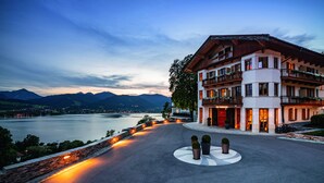 A hotel with a view of a lake and mountains. It has multiple balconies with plants and a stone wall along the lake. - Das Tegernsee (Tegernsee)