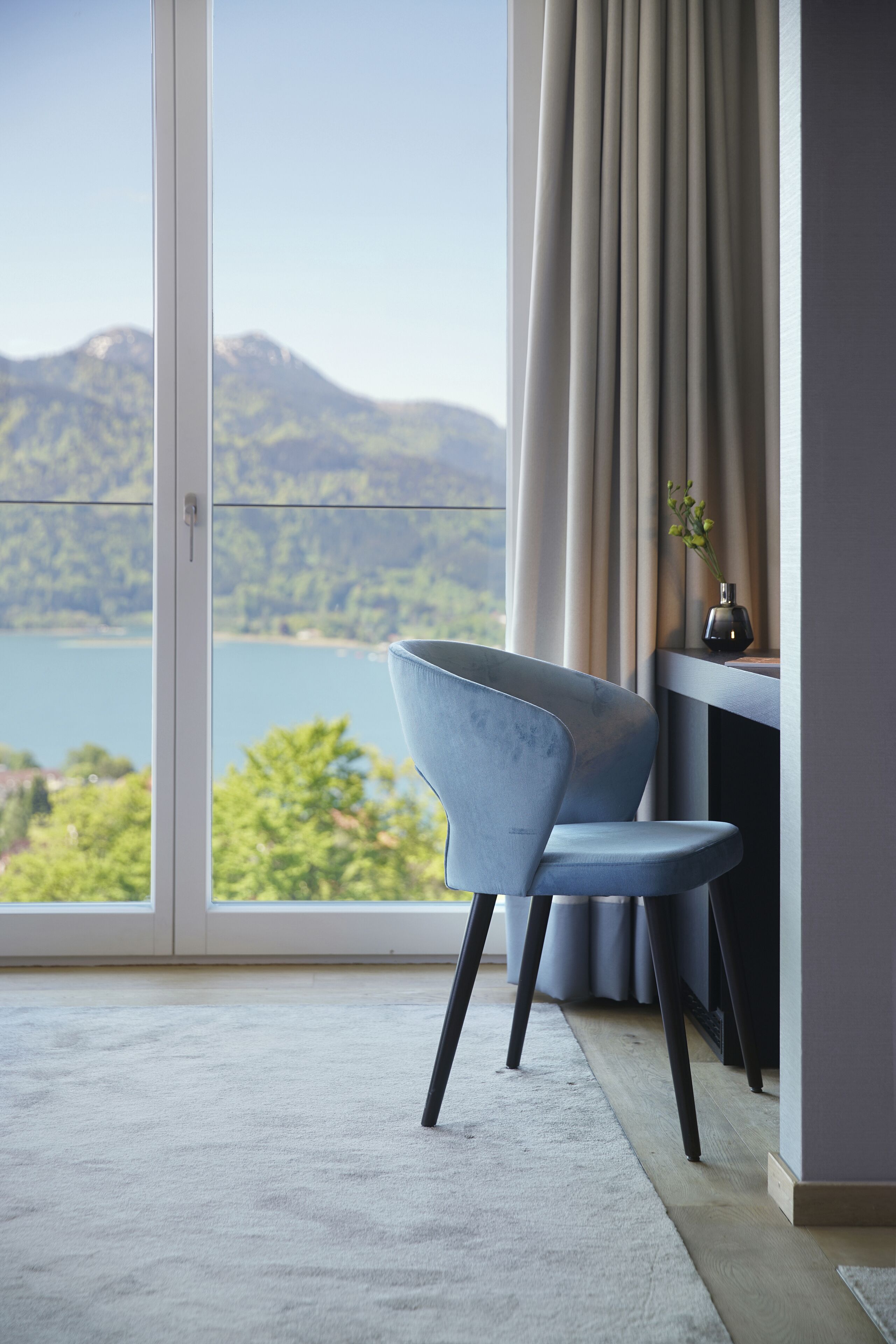 A modern room with a large window, a blue chair, a desk, and a vase with flowers.