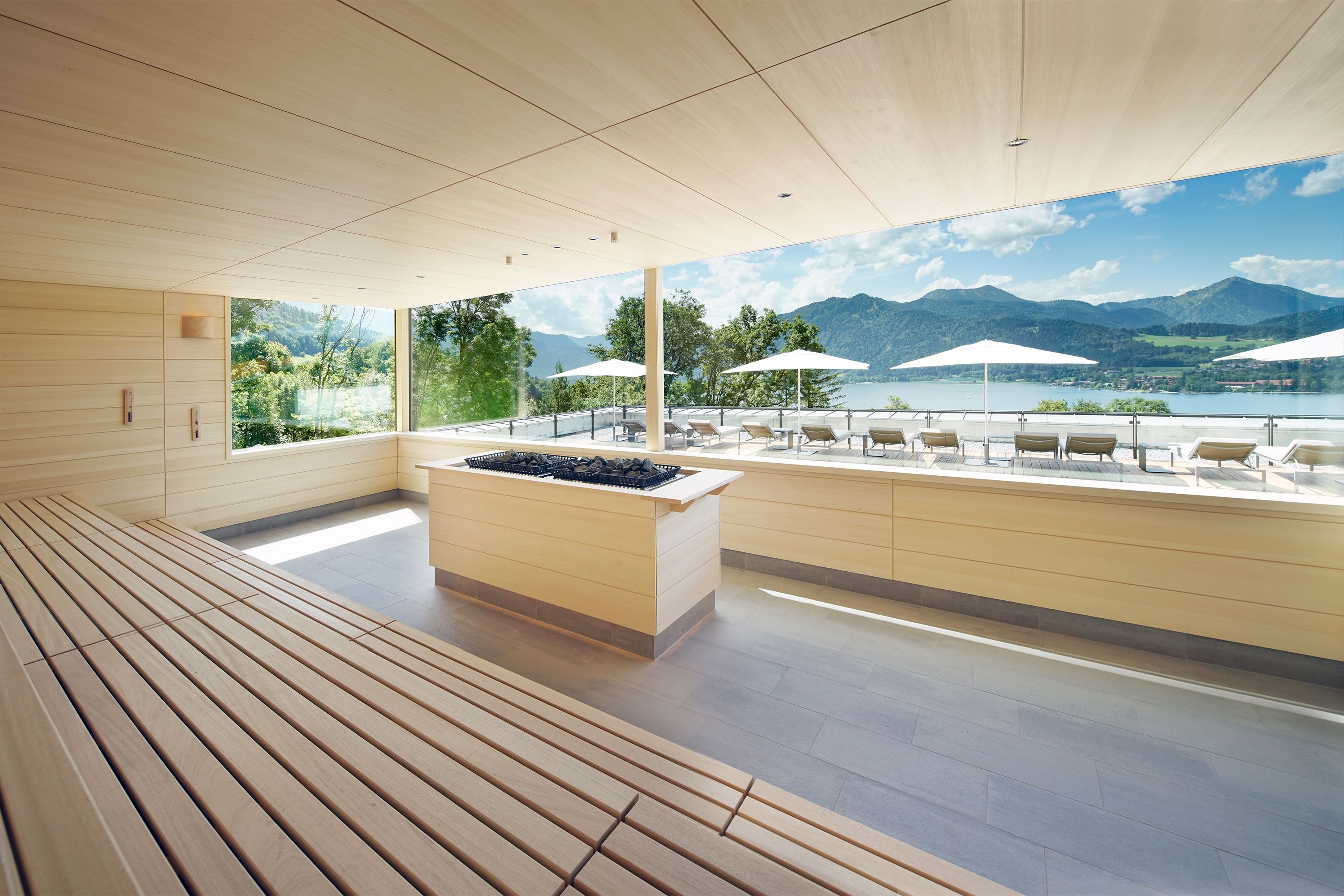 Un moderno espacio de spa con bancos de madera, una piscina climatizada en el centro y vistas a montañas y un lago.