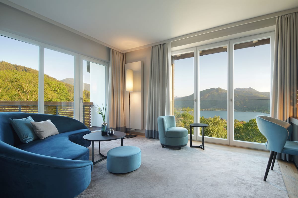 A modern room with a sofa, ottoman, and chairs, large windows with a view of a lake and mountains.