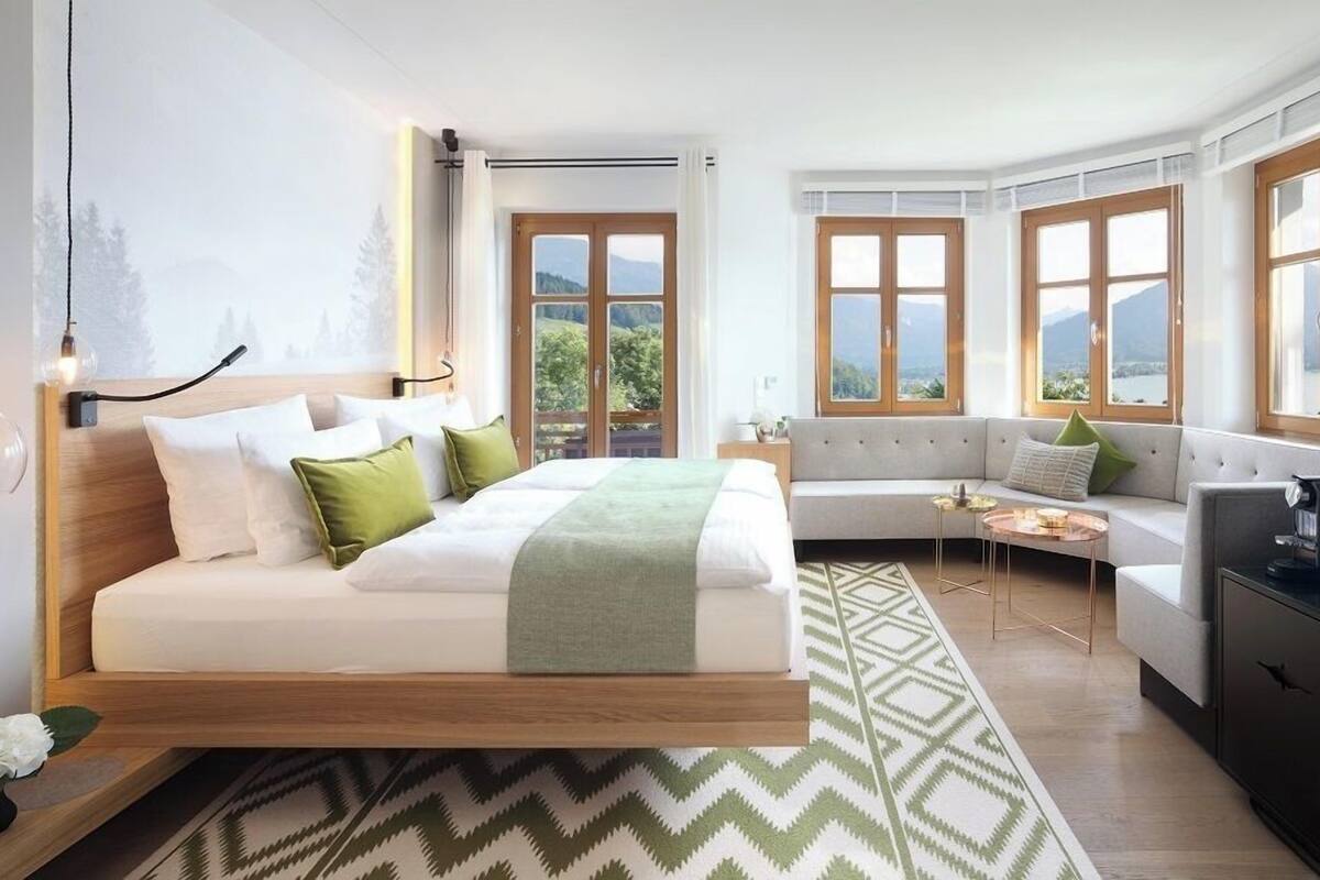 A modern bedroom with a bed, a sofa, and a desk. There are windows with a view of the outdoors.